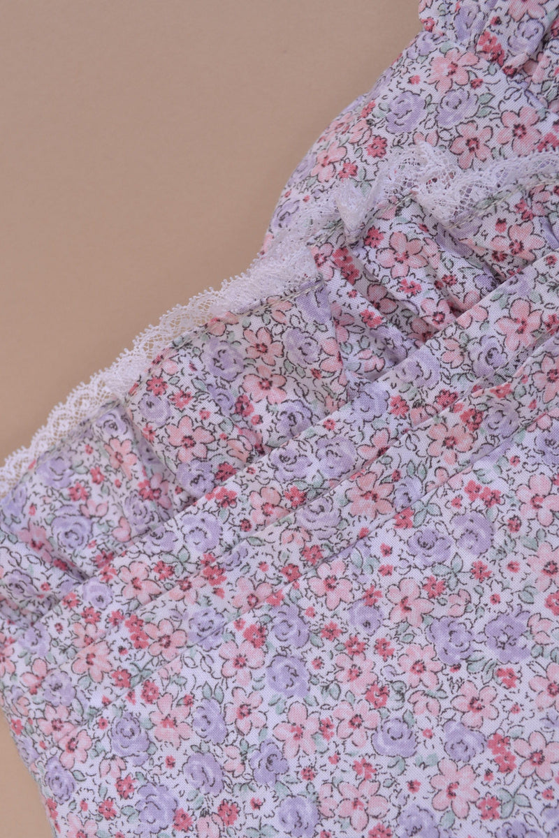 Capucine, Barboteuse fille, col rond, buste smocké, emmanchure volantée finition dentelle, 3 pressions sur le bas, en imprimé petites fleurs rose et lilas